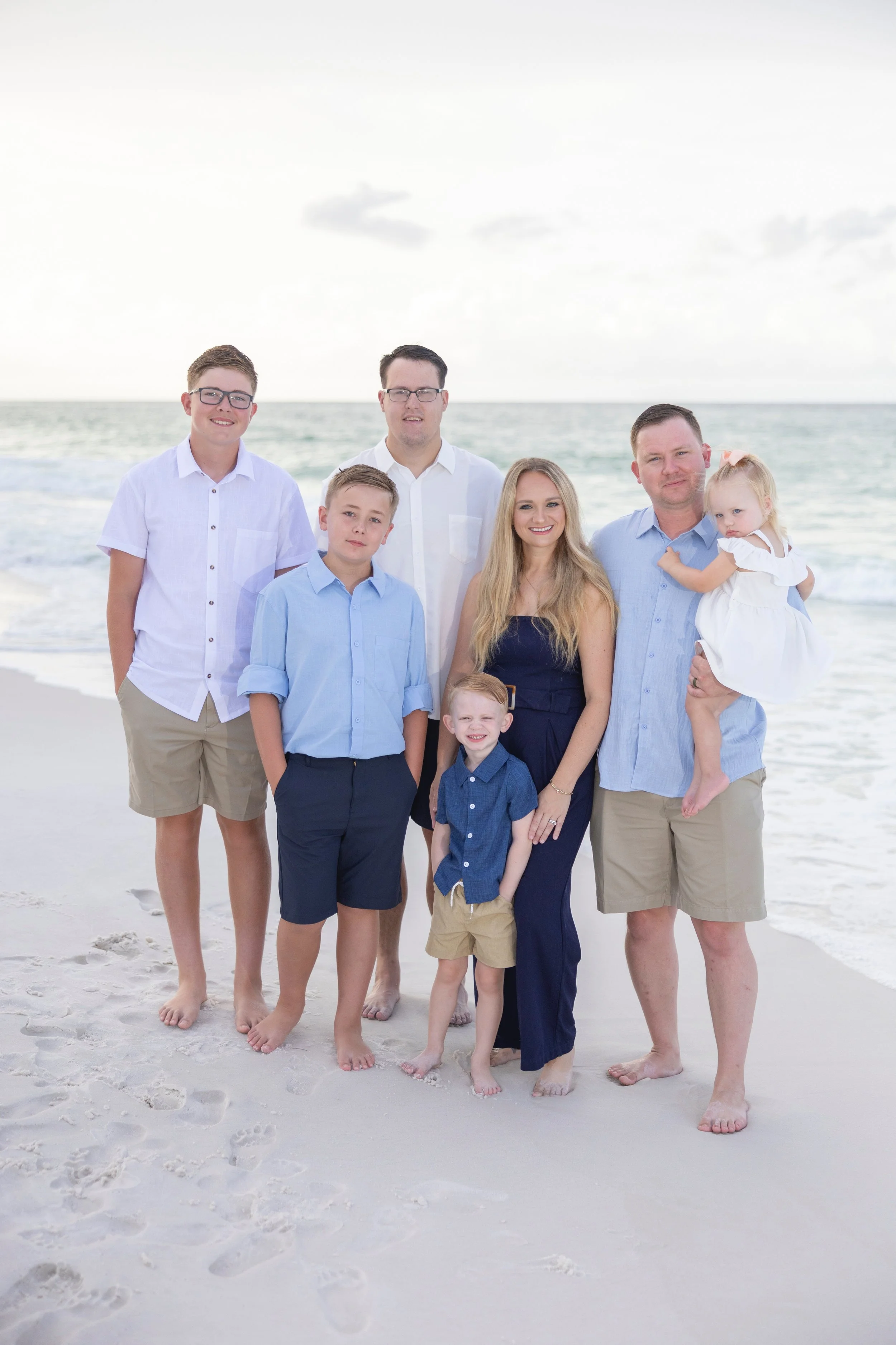 A family of seven standing on a sandy beach with the ocean behind them. The family includes three adult men and women and four children, all dressed in light-colored or navy blue clothing, with the adults smiling and the children posing for the camera.